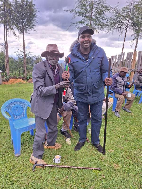 Long’ole in a blue jacket laughs beside an elder holding a beaded staff