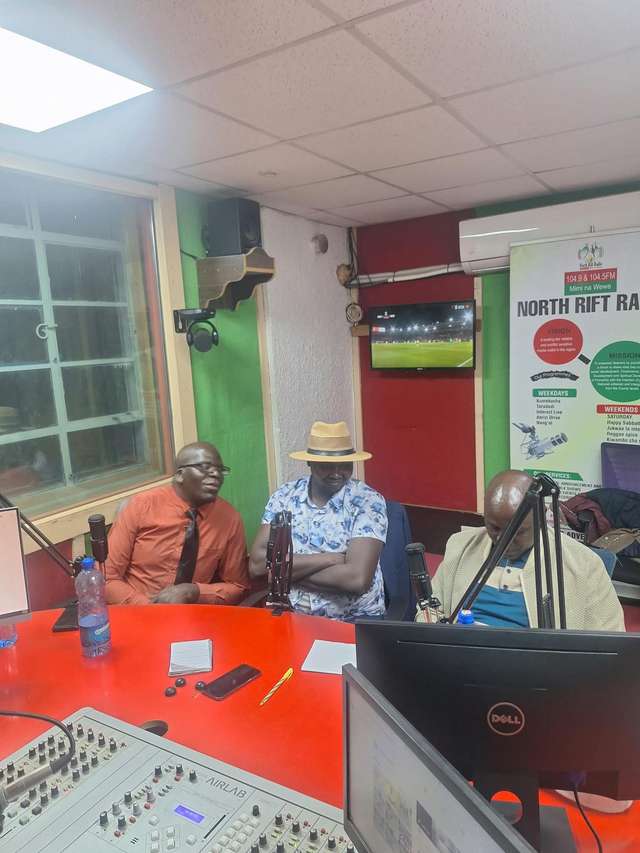 Long’ole in a hat at a radio studio table during a North Rift Radio interview